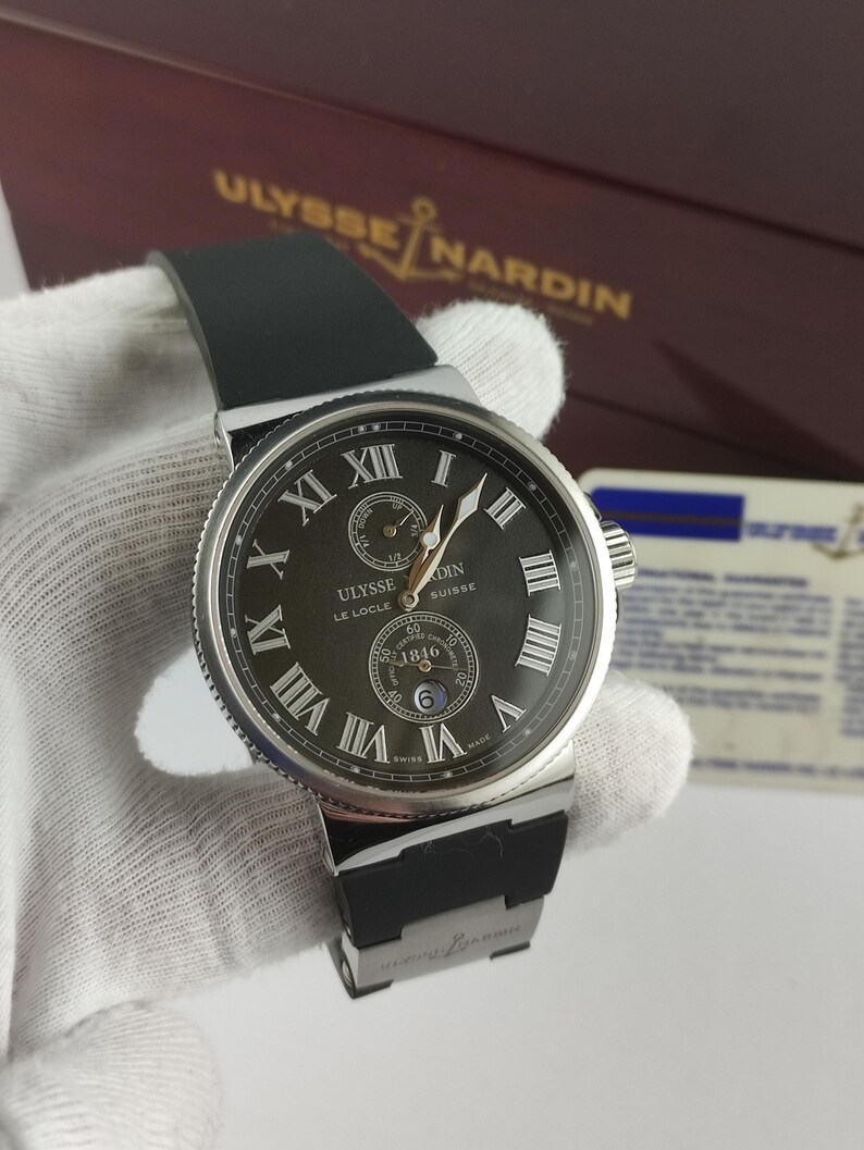 K&ouml;nnte beinhalten: Eine luxuri&ouml;se Armbanduhr mit schwarzem Zifferblatt, silberner L&uuml;nette und schwarzem Kautschukarmband. Die Uhr verf&uuml;gt &uuml;ber r&ouml;mische Ziffern und Hilfszifferbl&auml;tter. Der Markenname "Ulysse Nardin" ist auf dem Zifferblatt und der Box zu sehen.