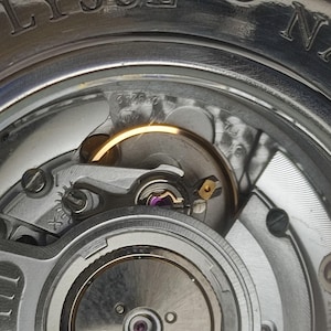 K&ouml;nnte beinhalten: Nahaufnahme eines Uhrwerks mit komplizierten Details. Das Bild zeigt die inneren Komponenten einer Uhr, einschlie&szlig;lich Zahnr&auml;dern, Schrauben und Unruh. Der Markenname ULYSSE NARDIN ist auf dem &auml;u&szlig;eren Geh&auml;use zu sehen.