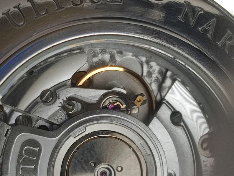 K&ouml;nnte beinhalten: Nahaufnahme eines Uhrwerks mit komplizierten Details. Das Bild zeigt die inneren Komponenten einer Uhr, einschlie&szlig;lich Zahnr&auml;dern, Schrauben und Unruh. Der Markenname ULYSSE NARDIN ist auf dem &auml;u&szlig;eren Geh&auml;use zu sehen.