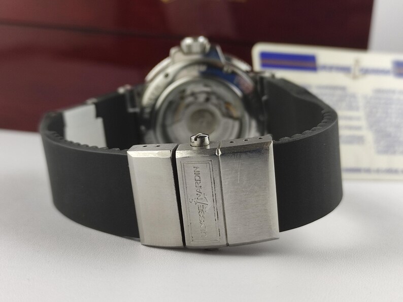 K&ouml;nnte beinhalten: Nahaufnahme einer Luxus-Armbanduhr mit schwarzem Kautschukarmband und silberner Schlie&szlig;e. Das Uhrwerk ist sichtbar. Die Schlie&szlig;e ist mit dem Markennamen "Ulysse Nardin" graviert.
