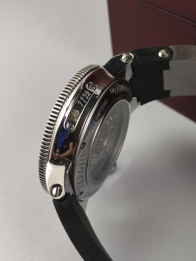 K&ouml;nnte beinhalten: Nahaufnahme einer silbernen Armbanduhr mit schwarzem Armband. Auf dem Zifferblatt steht "200 METERS" und "NARDIN". Die Uhr hat eine geriffelte L&uuml;nette und die Nummer "1132" ist sichtbar.