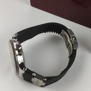 K&ouml;nnte beinhalten: Eine silberfarbene Armbanduhr mit schwarzem Kautschukband. Das Zifferblatt der Uhr ist rund mit einer strukturierten L&uuml;nette. Das Armband wird mit einem silbernen Verschluss befestigt. Die Uhr liegt auf einer wei&szlig;en Oberfl&auml;che.