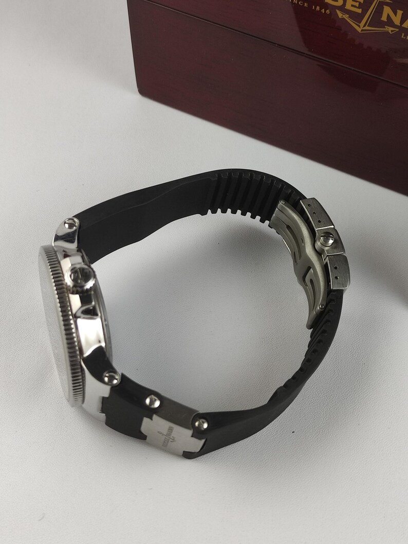 K&ouml;nnte beinhalten: Eine silberfarbene Armbanduhr mit schwarzem Kautschukband. Das Zifferblatt der Uhr ist rund mit einer strukturierten L&uuml;nette. Das Armband wird mit einem silbernen Verschluss befestigt. Die Uhr liegt auf einer wei&szlig;en Oberfl&auml;che.