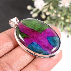 May include: An oval pendant featuring a polished, multi-coloured gemstone with vibrant patches of deep green, bright magenta, and electric blue set in a smooth, polished silver frame with a triangular bail at the top for a necklace chain.