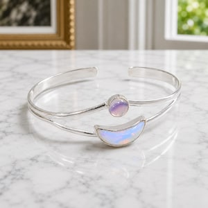 Puede incluir: Una pulsera brazalete de plata con dos bandas abiertas. Una banda sostiene un pequeño cabujón redondo iridiscente, mientras que la otra banda presenta una piedra iridiscente más grande en forma de media luna. El metal tiene un acabado pulido.