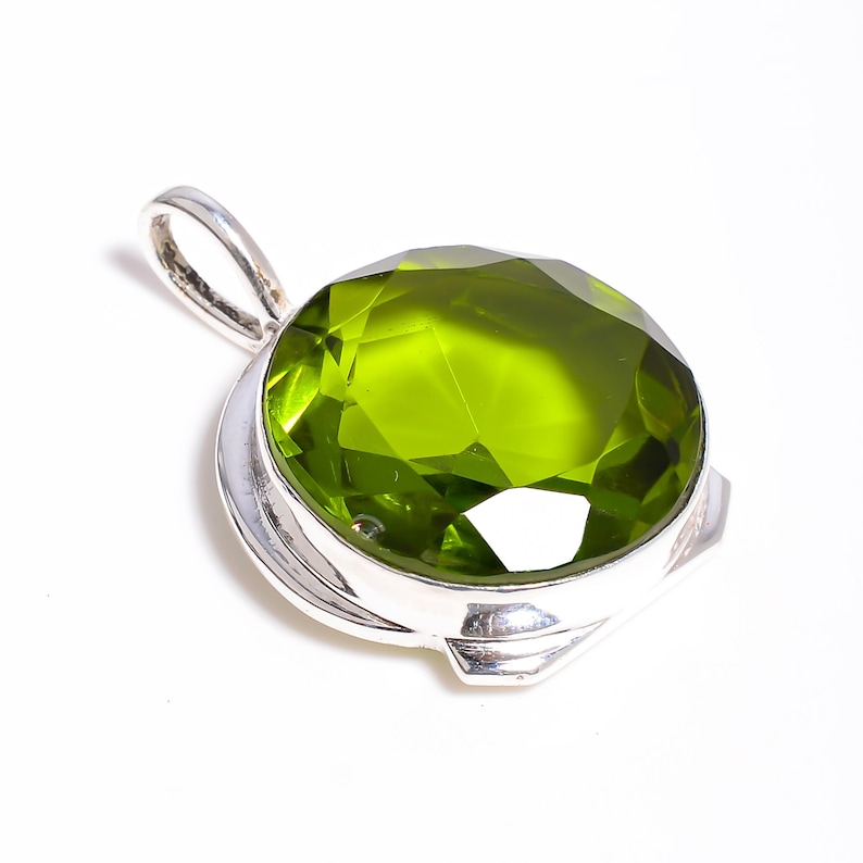 May include: A round, faceted, lime-green gemstone pendant set in a polished silver metal bezel. The pendant features a smooth, circular silver bail at the top for attachment, showcasing a vibrant, translucent green stone with multiple geometric facets.