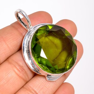May include: A round, faceted olive-green gemstone pendant set in a polished silver frame. The pendant features a large, circular stone with a brilliant cut, secured by a smooth silver bezel and topped with a sturdy, integrated silver bail loop.
