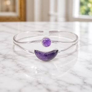 May include: A silver cuff bracelet featuring a polished, open-ended band design. The centre of the bracelet is adorned with a small, round, purple amethyst gemstone positioned above a larger, crescent-shaped, purple amethyst gemstone.