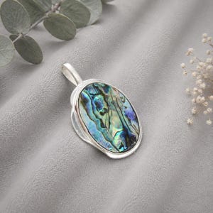 Könnte beinhalten: Ein ovaler Anhänger mit einer Einlage aus poliertem Abalone-Muschelgehäuse in irisierenden Blau-, Grün- und Violetttönen, eingefasst in einen glatten, silberfarbenen Metallrahmen mit einer stabilen Öse an der Oberseite für eine Halskette.