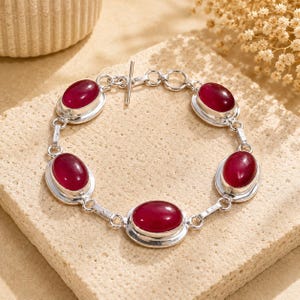 Può includere: Un bracciale a maglie in argento con cinque pietre preziose rosso scuro di forma ovale, incastonate in castoni d'argento lucido. Il bracciale è fissato con una chiusura a T in argento, composta da un anello circolare e una barretta.
