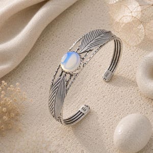 Peut inclure: Un bracelet manchette en argent orné d'un cabochon central rond en pierre de lune irisée, encadré par deux motifs de plumes gravés. La bande métallique présente une finition polie avec des détails oxydés et porte le poinçon 925 à l'intérieur.