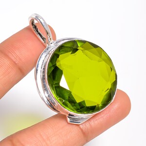 May include: A round, faceted lime green gemstone pendant set in a polished silver metal frame. The pendant features a prominent bail at the top for attachment to a chain, showcasing a vibrant, translucent green stone with a circular cut.