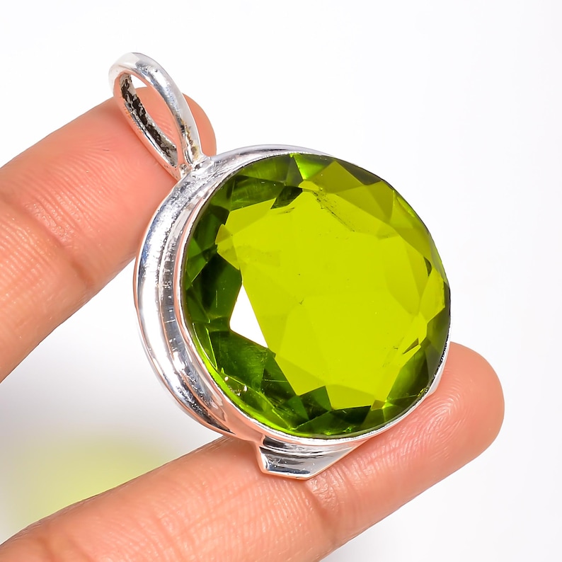 May include: A round, faceted lime green gemstone pendant set in a polished silver metal frame. The pendant features a prominent bail at the top for attachment to a chain, showcasing a vibrant, translucent green stone with a circular cut.
