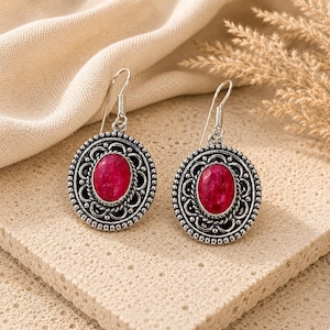 Peut inclure: Une paire de boucles d'oreilles pendantes argentées avec une pierre rose vif de forme ovale au centre. La monture en métal présente une bordure texturée complexe avec un contour perlé et une attache en crochet pour l'oreille.