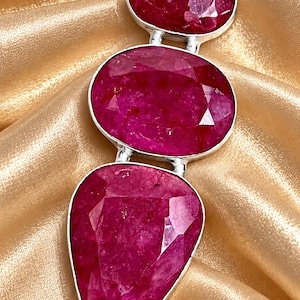 May include: A pendant featuring three faceted, deep pinkish-red ruby gemstones set in silver. The stones are arranged vertically, with a small oval at the top, a larger oval in the center, and a pear-shaped stone at the bottom, all linked together.