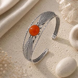 Peut inclure: Un bracelet ouvert en métal argenté oxydé, orné de deux motifs symétriques en forme de feuille et d'une pierre orange ronde et lisse en son centre. Le bracelet est composé de plusieurs fils fins parallèles et porte le poinçon 925 sur le métal.