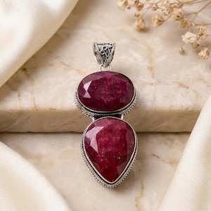 May include: A pendant featuring two faceted, deep red stones set in silver. The top stone is oval-shaped, while the bottom stone is pear-shaped. The silver setting includes a decorative, filigree-style bail at the top for attaching to a chain.