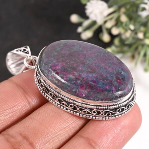 Puede incluir: Un colgante ovalado con una piedra preciosa de rubí zoisita pulida con patrones moteados en magenta intenso y verde oscuro. La piedra está engastada en un marco decorativo de plata de ley con filigrana intrincada y una anilla a juego.