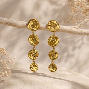 Puede incluir: Un par de pendientes colgantes de tono dorado, cada uno con cuatro discos circulares de textura martillada unidos verticalmente en orden de tamaño decreciente. La superficie metálica tiene un acabado reflectante e irregular con formas orgánicas.