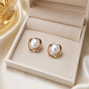 Puede incluir: Un par de pendientes de botón con una perla esférica blanca central rodeada por un marco de metal dorado de forma irregular y diseño abierto. Los pendientes se presentan dentro de un joyero de color claro forrado en terciopelo con tapa.