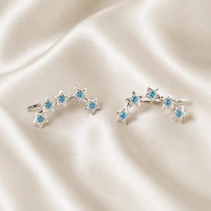 May include: A pair of silver-toned ear climbers, each featuring a curved row of four five-pointed stars. Each star is set with a small, round, bright blue gemstone in the center, creating a celestial design that follows the curve of the ear.
