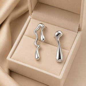 May include: A pair of silver-toned earrings displayed in a beige jewelry box. One earring has a flowing, asymmetrical design, while the other is a more streamlined, elongated shape. The earrings have a polished, reflective surface.
