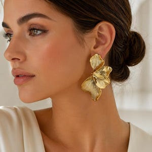 May include: Gold-colored statement earrings in a floral design. The earrings feature three overlapping petals with textured detailing. The earrings are worn on the ear of a person with dark hair and a white top.