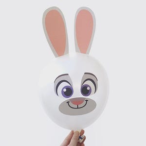 May include: A white balloon with a cartoon rabbit face, featuring large purple eyes, a gray nose, and a smiling mouth. The balloon has large, pink-lined ears. The balloon is held by a hand against a white background.