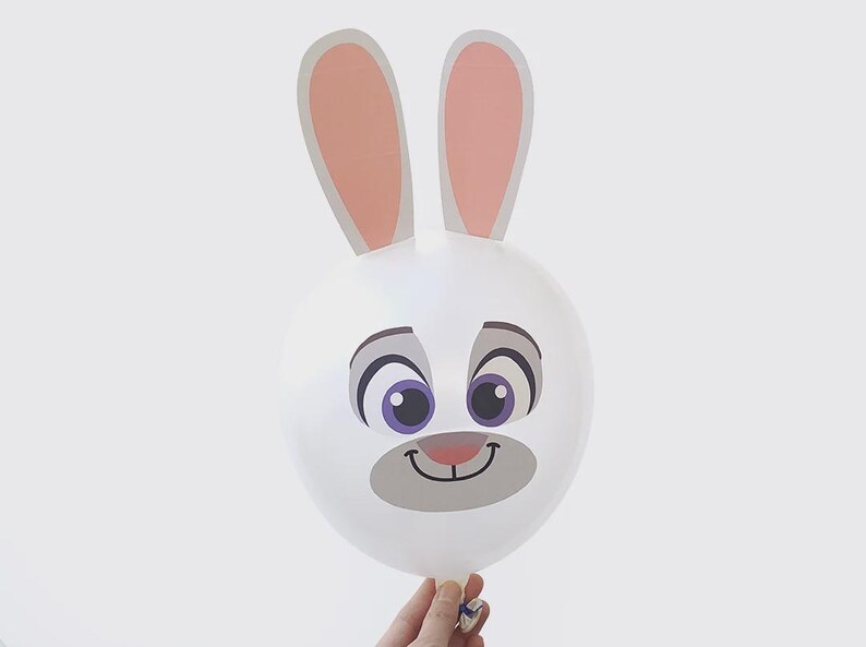 May include: A white balloon with a cartoon rabbit face, featuring large purple eyes, a gray nose, and a smiling mouth. The balloon has large, pink-lined ears. The balloon is held by a hand against a white background.