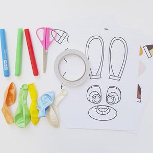 May include: A collection of craft supplies for a DIY project, including blue, green, and red markers, pink scissors, tape, deflated balloons, and printed bunny face and ear templates on white paper. The supplies are arranged on a white surface.