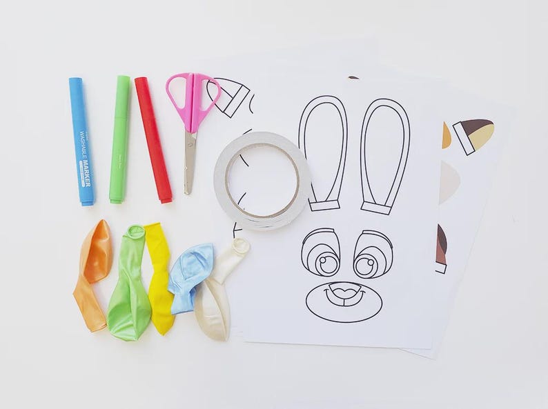 May include: A collection of craft supplies for a DIY project, including blue, green, and red markers, pink scissors, tape, deflated balloons, and printed bunny face and ear templates on white paper. The supplies are arranged on a white surface.