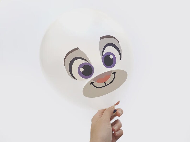 May include: A white balloon with a cartoon face printed on it. The face has large eyes with purple irises, gray eyebrows, and a smiling mouth. The balloon is held by a hand against a white background.