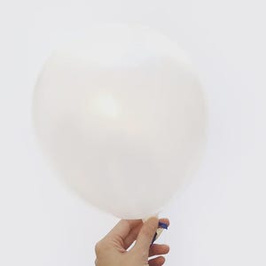 May include: A white balloon, held by a hand, against a white background. The balloon is inflated and appears to be made of a translucent material. A small piece of blue tape secures the balloon's neck.