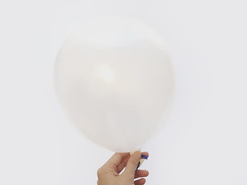 May include: A white balloon, held by a hand, against a white background. The balloon is inflated and appears to be made of a translucent material. A small piece of blue tape secures the balloon's neck.
