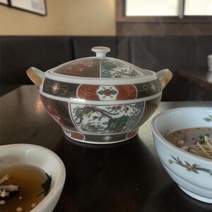 Peut inclure: Une soupière décorative en céramique avec un couvercle, ornée d'un motif floral et d'oiseaux colorés. La soupière a des poignées dorées et est posée sur une table en bois sombre. Un bol de soupe est visible en arrière-plan.