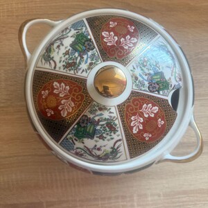 Peut inclure: Un plat de service rond avec couvercle, poignées dorées et bouton doré. Le couvercle présente un motif décoratif avec des motifs floraux et géométriques rouges, verts et blancs. Le plat a un bord noir et un intérieur blanc.