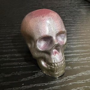 Puede incluir: Una escultura decorativa de calavera con acabado metálico en tonos plateados, morados y rosas. La calavera tiene rasgos detallados, incluyendo cuencas de los ojos, cavidad nasal y dientes. La superficie tiene una calidad brillante e iridiscente.