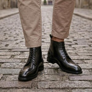 May include: A pair of black leather ankle boots with a polished finish, featuring a rounded toe, a lace-up front with thin black laces, and a low heel, worn with beige trousers that end just above the ankle to showcase the boot silhouette.