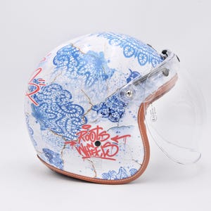 Könnte beinhalten: Weißer Motorradhelm mit blauen Blumenmustern und rotem Schriftzug "Roots Wheel". Mit einem transparenten Visier und braunem Lederbesatz. Ein stilvolles und schützendes Accessoire.