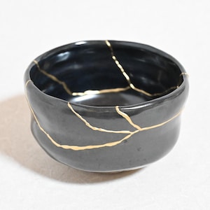 Può includere: Una piccola ciotola nera rotonda con una finitura lucida e linee irregolari color oro che attraversano la superficie, collegando varie sezioni della ciotola in un motivo che ricorda le tecniche di riparazione della ceramica.