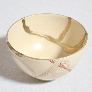Op de afbeelding: Crèmekleurige keramische kom met goudkleurige kintsugi-reparatielijnen. De kom heeft een afgeronde vorm en een decoratief patroon van rode en beige lijnen bij de basis. Een uniek stuk aardewerk.