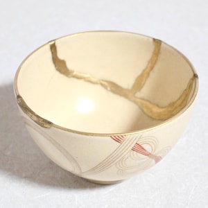 May include: Cream-colored ceramic bowl with gold-colored kintsugi repair lines. The bowl has a rounded shape with a decorative pattern of red and beige lines near the base. The kintsugi technique highlights the history of the bowl.
