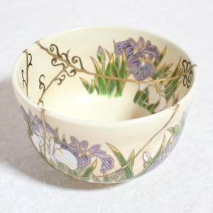 May include: Cream-colored ceramic bowl with a floral design featuring purple irises and green leaves. The bowl has gold accents and visible repair lines. The design wraps around the exterior of the bowl.