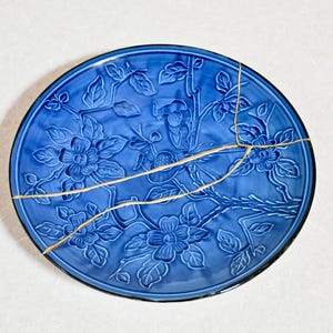 May include: A circular blue ceramic plate featuring a raised relief pattern of flowers and branches. The plate is repaired with gold-colored lacquer lines across the surface, following the kintsugi technique, highlighting the cracks in the ceramic.
