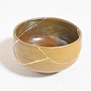 May include: A round, ceramic bowl with a warm, earthy color palette. The bowl has been repaired using the Kintsugi method, with gold-colored lines highlighting the cracks. The interior is a darker brown, contrasting with the lighter exterior.