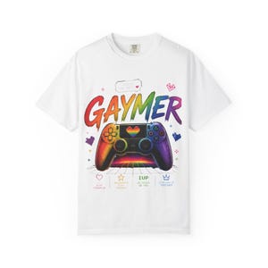 May include: White t-shirt with a colorful graphic of a video game controller in rainbow colors. The word "GAYMER" is written in large, vibrant letters above the controller. Additional text includes "Pause the hate" and other gaming-related phrases.