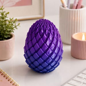 May include: An egg-shaped ornament with a geometric design, transitioning from purple to blue. The object is displayed on a white surface, possibly a desk or table. The colour gradient and pattern create a striking visual effect.