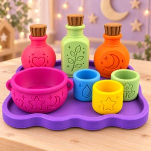 May include: A set of colorful silicone play items on a purple tray, including a pink cauldron with star and moon motifs, three bottles with brown corks, and three small cups featuring embossed icons like a water drop, flower, and star.