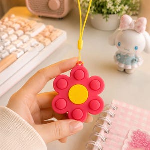May include: A pink flower-shaped fidget toy with a yellow center and raised pink circles. It has a yellow lanyard attached. The toy is held in a hand, with other desk items in the background.
