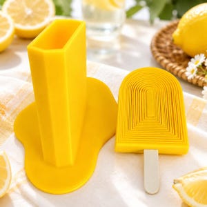 May include: Two bright yellow ice lolly moulds. One is a rectangular shape, the other is a layered design with a white stick. The rectangular mould has a base that resembles a puddle. Lemons and a woven basket are in the background.
